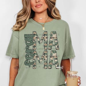 Op de afbeelding: Saliegroen t-shirt met de woorden "Ferall Mama" in camouflageprint. Het shirt heeft een relaxte pasvorm en korte mouwen. De tekst is zwart omlijnd en voorzien van een sterrenontwerp.