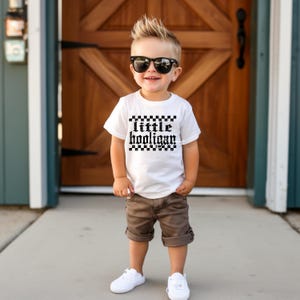 K&ouml;nnte beinhalten: Ein kleiner Junge tr&auml;gt ein wei&szlig;es T-Shirt mit einem schwarz-wei&szlig;en Karomuster und dem Text "little hooligan" darauf. Er tr&auml;gt auch braune Shorts, wei&szlig;e Turnschuhe und eine Sonnenbrille.