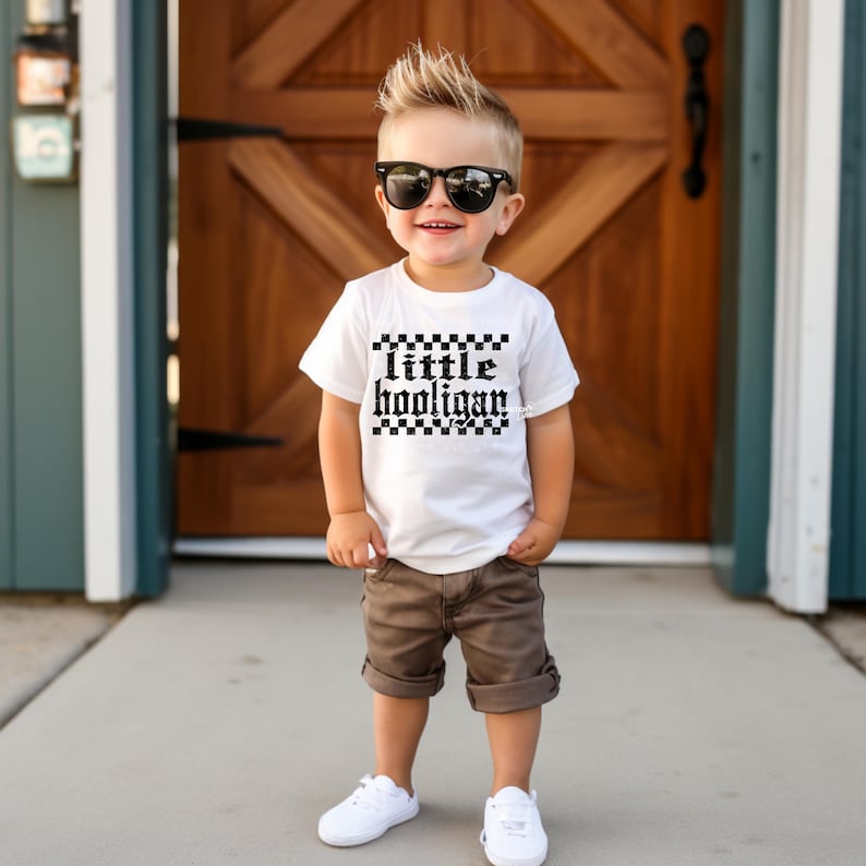 K&ouml;nnte beinhalten: Ein kleiner Junge tr&auml;gt ein wei&szlig;es T-Shirt mit einem schwarz-wei&szlig;en Karomuster und dem Text "little hooligan" darauf. Er tr&auml;gt auch braune Shorts, wei&szlig;e Turnschuhe und eine Sonnenbrille.