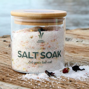 May include: A glass jar with a bamboo lid filled with pink Himalayan salt and Epsom salt. The label reads "Morro Mermaids Pacific Sea Epsom Salt Soak Let your tail out 18oz."