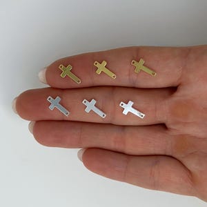 May include: Six small cross charms in gold and silver tones are displayed. The charms are arranged on a person's fingers, with three gold crosses on top and three silver crosses below. The crosses have a simple, minimalist design.