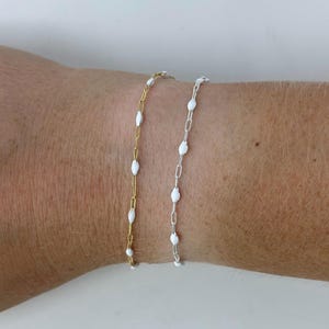 Könnte beinhalten: Zwei zarte Armbänder, eines goldfarben und eines silberfarben, jeweils mit kleinen, weißen, oval geformten Perlen verziert. Die Armbänder werden an einem Handgelenk vor einem schlichten weißen Hintergrund präsentiert, was ihr minimalistisches Design und ihre elegante Schlichtheit hervorhebt.