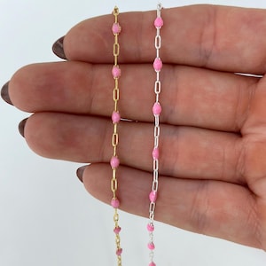 May include: Two delicate necklaces with pink bead accents. One necklace is gold-toned, the other is silver-toned. The chains are held against a neutral background, showcasing the jewelry's design and color.