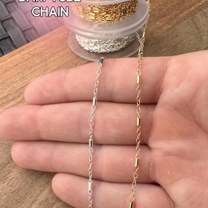 May include: Close-up of two bar/tube chains, one silver and one gold, displayed against a wooden surface. The chains are made of small, rectangular bars connected by delicate links. The text "BAR/TUBE CHAIN" is visible in the upper left corner.