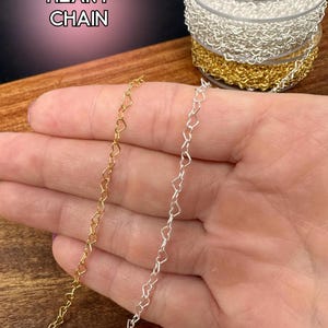 May include: Gold and silver heart chain displayed on a wooden surface. The image shows two strands of chain, one gold and one silver, each with heart-shaped links. The text "HEART CHAIN" is visible in the upper left corner.