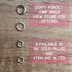 May include: Four silver jump rings of varying sizes are displayed on a wooden surface. Text on a pink background reads "DON'T FORGET JUMP RINGS! VIEW STORE FOR OPTIONS!" and "AVAILABLE IN 14K GOLD FILLED AND STERLING SILVER."