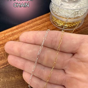 May include: Silver and gold paperclip chain displayed on a wooden surface. The image shows two chains, one silver and one gold, with an elongated oval link design. The text "PAPERCLIP CHAIN" is in the upper left corner.