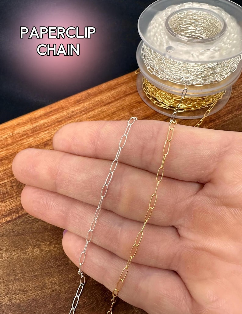 May include: Silver and gold paperclip chain displayed on a wooden surface. The image shows two chains, one silver and one gold, with an elongated oval link design. The text "PAPERCLIP CHAIN" is in the upper left corner.