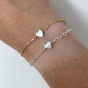 May include: Two delicate bracelets, one gold-toned and one silver-toned, each featuring a small, white heart charm. The gold bracelet has a chain-link design, while the silver bracelet has a delicate chain. The bracelets are worn on a wrist.