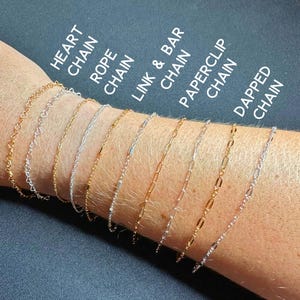 May include: A selection of delicate chain bracelets in gold and silver, displayed on a wrist. Styles include heart, rope, link & bar, paperclip, and dapped chains. Text labels identify each style. The background is a dark, solid color.