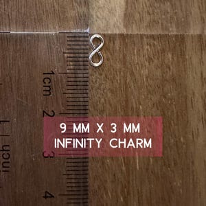 May include: A silver infinity charm measuring 9 mm by 3 mm. The charm is shown on a wooden surface with a ruler for scale.