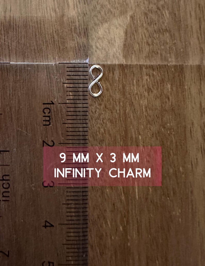 May include: A silver infinity charm measuring 9 mm by 3 mm. The charm is shown on a wooden surface with a ruler for scale.