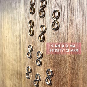 May include: Ten gold-colored infinity charm pendants, each measuring 9 mm by 3 mm. The charms are arranged in a pattern on a wooden surface.