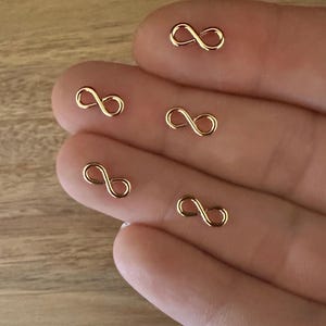 May include: Five gold infinity symbol charms on a hand. The charms are small and delicate.