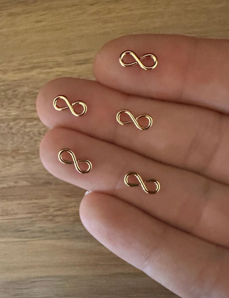 May include: Five gold infinity symbol charms on a hand. The charms are small and delicate.
