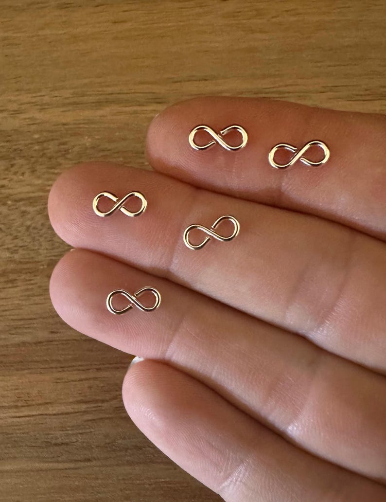 May include: Five silver infinity symbol charms on a hand. The charms are small and delicate.