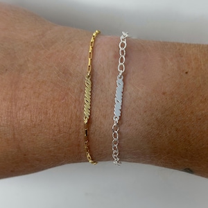 May include: Two bracelets, one gold and one silver, each with the word "mama" in cursive script. The gold bracelet has a rectangular link chain, the silver one a chain link design. Both are shown on a wrist. The bracelets are likely made of metal.