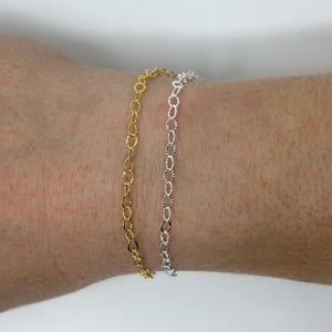 May include: Two chain bracelets, one gold and one silver, on a person's wrist. The bracelets are made of a delicate, oval-shaped chain.
