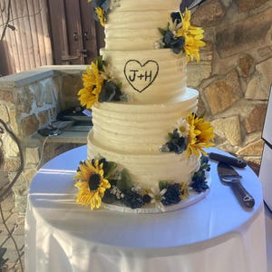 May include: A three-tiered wedding cake with white frosting and sunflower decorations. The cake has a heart with the initials "J+H" drawn on the front. The cake is on a white tablecloth.