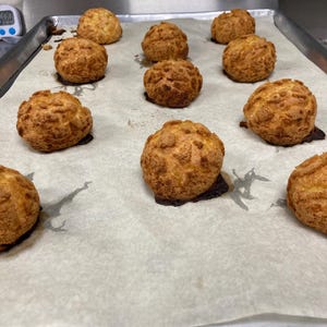 Pode incluir: Uma assadeira com papel vegetal contém uma dúzia de profiteroles dourados. Os produtos assados ​​têm uma superfície texturizada e enrugada e são dispostos em fileiras. A assadeira está em um ambiente de cozinha comercial.