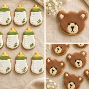 May include: A collection of decorated sugar cookies featuring white baby bottles with green and yellow icing details, alongside brown bear face cookies with cream-colored muzzles, black eyes, and noses, arranged on a neutral fabric surface.