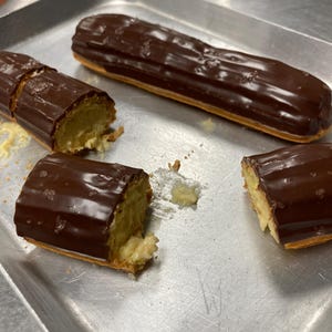 Pode incluir: Vários éclairs de chocolate numa bandeja de metal. Um éclair inteiro e dois cortados em pedaços, revelando um recheio cremoso. Os éclairs são cobertos com uma cobertura de chocolate preto brilhante. Uma sobremesa deliciosa.
