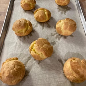 Pode incluir: Uma assadeira com oito profiteroles dourados, dispostos sobre papel vegetal. Os profiteroles têm uma forma arredondada com uma superfície ligeiramente texturizada. O fundo mostra uma superfície de madeira e um forno de metal.
