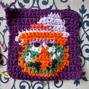 May include: Crocheted square with a purple background and a colourful pumpkin face in the centre. The pumpkin has a white hat with a pink brim and black eyes.