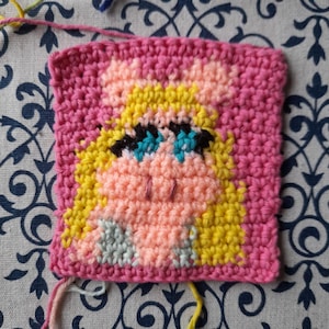 May include: A crocheted square featuring a cartoon character with blonde hair, blue eyes, and a pink face. The character is surrounded by a pink border.