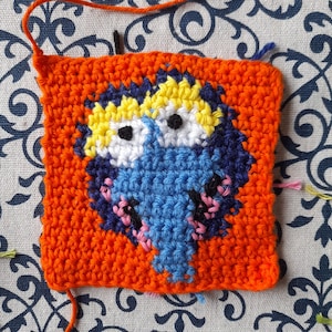 May include: A crocheted square with a blue, white, yellow, and pink design. The square is surrounded by a bright orange border.