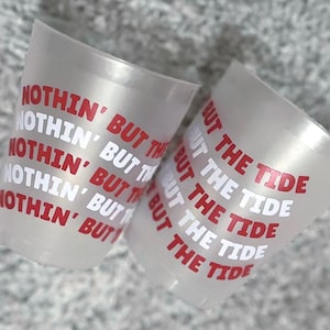 May include: Two clear plastic shot glasses with the text "NOTHIN' BUT THE TIDE" in red and white. The glasses are slightly translucent and angled towards the viewer, set against a textured gray background.