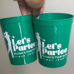 Pode incluir: Dois copos de plástico verde com texto branco que diz "Let's Partee - Elijah's Thirtieth - est 1995". Os copos apresentam uma silhueta de um golfista.
