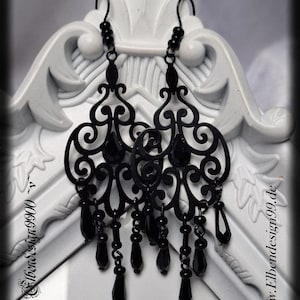 Peut inclure: Boucles d'oreilles noires en forme de lustre avec un design filigrané complexe et des perles en forme de goutte suspendues. Les boucles d'oreilles ont une fermeture à crochet et sont présentées sur un fond décoratif blanc. Le site web www.Elbendesign99.de est visible.