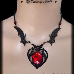May include: A black gothic necklace with bat wing details and a heart-shaped pendant holding a large, faceted red gemstone. The necklace is on a mannequin. The website address www.Elbendesign99.de is visible. The necklace has a delicate chain.