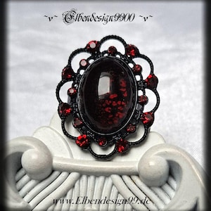 Peut inclure: Une bague en métal noir avec une pièce centrale ovale incrustée de taches rouge foncé. Le sertissage est entouré d'un cadre festonné orné de petites pierres rouges rondes à facettes. Le texte Elbendesign9900 et www.Elbendesign99.de est visible.