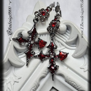 以下が含まれることがあります： 十字架のデザイン、赤い宝石のアクセント、ハート型の宝石が特徴のゴシック様式のイヤリング。 イヤリングは暗い金属仕上げで、白い装飾的な表面に表示されています。 www.Elbendesign99.deのウェブサイトアドレスが表示されています。