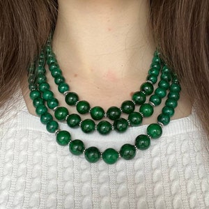 May include: A multi-strand necklace featuring three layers of polished, deep green spherical beads. Each green bead is separated by small, textured silver-toned metal spacers, creating a consistent pattern throughout the three strands of the necklace.