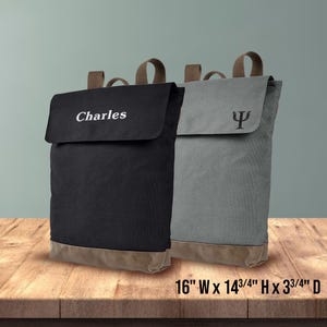 May include: Two canvas backpacks, one black and one gray, with brown accents. The black backpack has the name "Charles" embroidered on the flap. The gray backpack features the Greek letter psi. Each measures 16" W x 14 3/4" H x 3 3/4" D.
