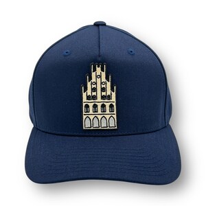 Könnte beinhalten: Marineblaue Baseballkappe mit einem gestickten Design eines Gebäudes mit mehreren Fenstern und einem Turm. Die Kappe hat einen gebogenen Schirm und eine strukturierte Krone. Das gestickte Design ist in Beige und Schwarz gehalten.
