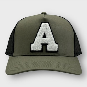 Könnte beinhalten: Eine grüne und schwarze Trucker-Kappe mit einem weißen Filzbuchstaben 'A' auf der Vorderseite.