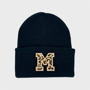 Könnte beinhalten: Eine marineblaue Strickmütze mit umgeschlagenem Saum. Die Mütze hat einen großen, gestickten Buchstaben "M" mit Leopardenmuster. Ein Winteraccessoire.