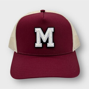 Könnte beinhalten: Eine burgunderrote und beige Trucker-Kappe mit einem weißen Filz-Buchstaben "M" auf der Vorderseite.