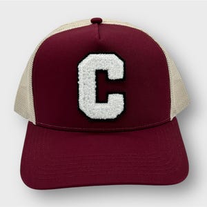 Könnte beinhalten: Eine burgunderrote und beige Trucker-Kappe mit einem weißen Filzbuchstaben 'C' auf der Vorderseite.