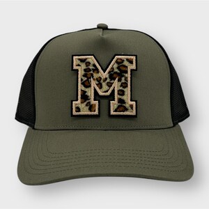 Könnte beinhalten: Olivgrüne Trucker-Cap mit schwarzem Mesh-Rücken und gebogenem Schirm. Auf der Vorderseite befindet sich ein großer, gestickter Buchstabe "M" im Leopardenmuster mit beigem Rand. Die Kappe hat einen klassischen, lässigen Stil.