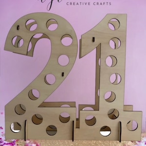May include: Wooden number 21 with holes for holding drinks. The number is made of light brown wood and is standing on a table with confetti.