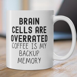 Op de afbeelding: Een witte keramische koffiemok met zwarte tekst die "Brain cells are overrated coffee is my backup memory" luidt.