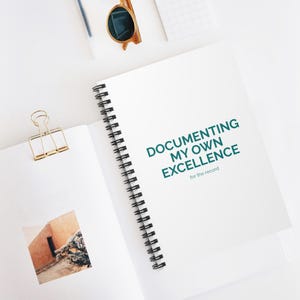 May include: A white spiral-bound notebook with the text "DOCUMENTING MY OWN EXCELLENCE for the record" in teal. The notebook features a gold metal binder clip on the left page, which displays a small square photograph of a desert building.