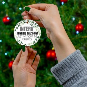 May include: A round white Christmas ornament with the text "INTERN RUNNING THE SHOW JUST WITHOUT THE PAYCHECK" in black. The ornament is decorated with green leaves and is held by a person in front of a Christmas tree.