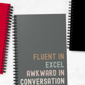 May include: A gray spiral bound notebook with the text "Fluent in Excel, Awkward in Conversation" printed in colorful letters.