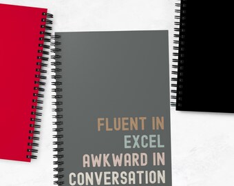 Könnte beinhalten: Ein graues Spiralblock-Notizbuch mit dem Text "Fluent in Excel, Awkward in Conversation", der in bunten Buchstaben gedruckt ist.
